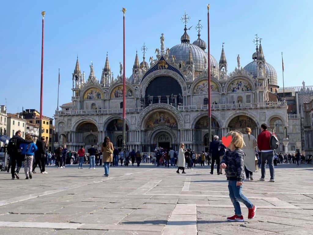 Child walks in front of the Basilica di San Marco in Venice, in the main piazza. Others walk around. They're wearing long sleeves or jackets.