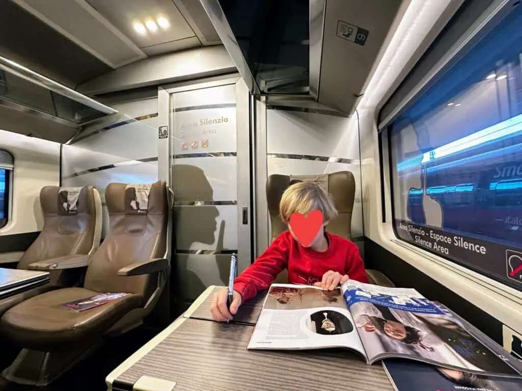 Boy holds pen and looks at magazine in seated area of Frecciarossa train in Italy.