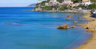 People swim and walk on the beach. Rocks in the water, buildings on a promontory in background.