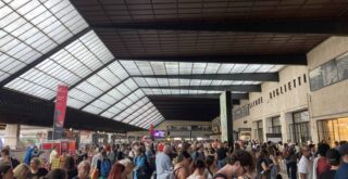 Glass roof over the Florence Santa Maria Novella train station. Crowd of people waits in the main area of the station looking up at the departures board on the right. To the left are the entrances to the tracks.