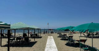 White walkway leads to beach and sea at the beach in Tuscany. Green and green and white striped umbrellas and tents are on either side.