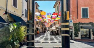 Enormous frame frames a street in Pietrasanta, Italy. There are colorful umbrellas hanging across the street.