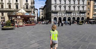 Boy in Semenyo soccer jersey walks in the middle of Piazza della Repubblica in Florence. Historic carousel ahead of him on left and multi-story buildings ahead of him. Others walk in the far end of the piazza.