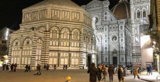 The Baptistry and the Duomo of Florence, Italy at night. They are both lit up and there are a few people walking in the square.